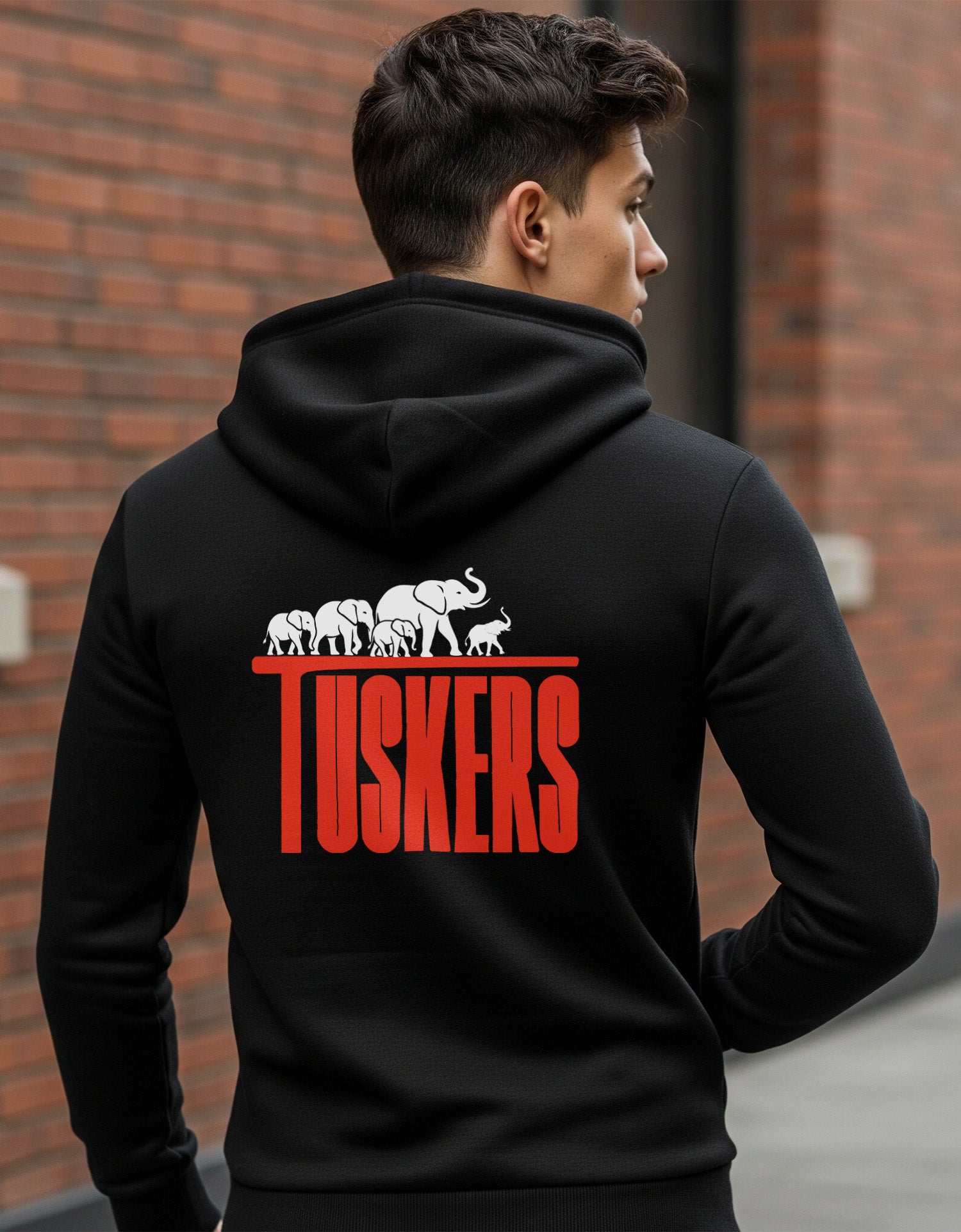 Person wearing a Dumkee black hoodie with 'Tuskers' design on the back against a brick wall.