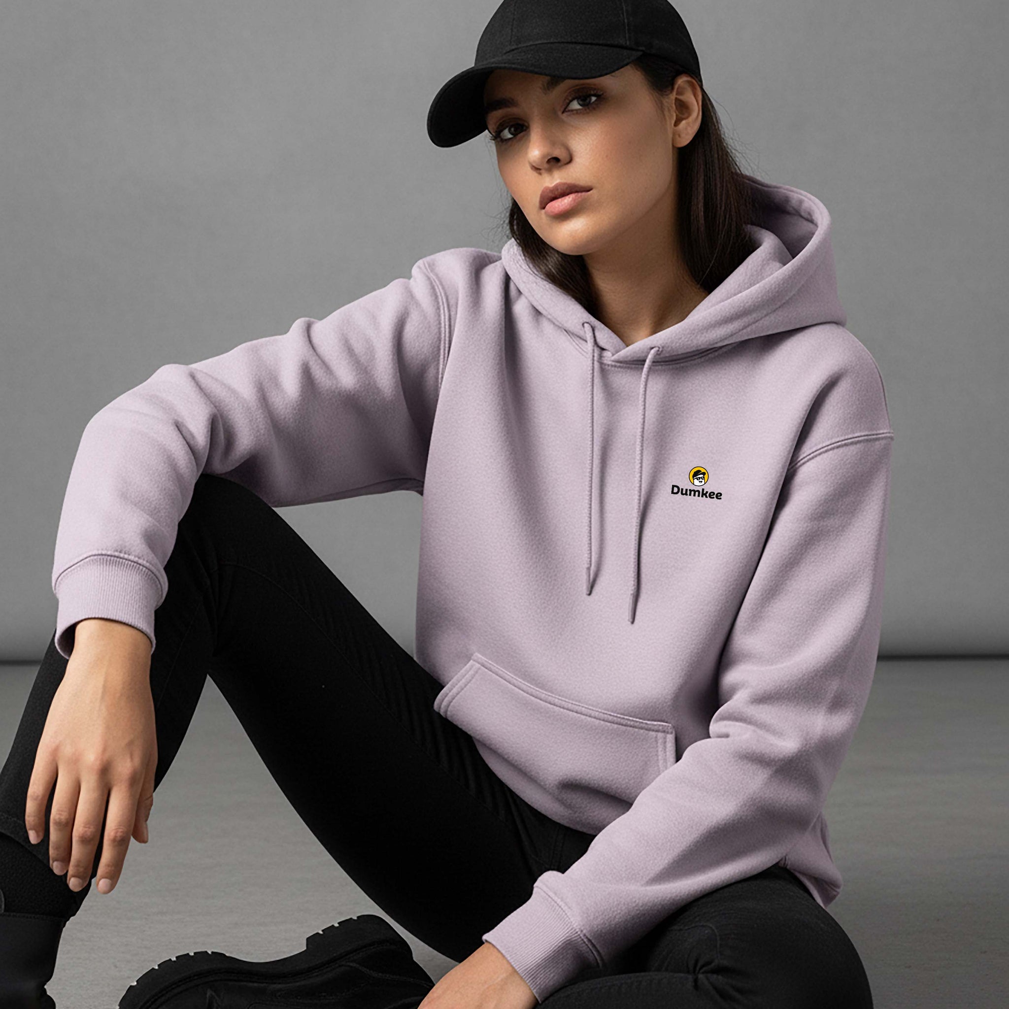 Person wearing a Dumkee baby pink hoodie with a logo, sitting on a gray floor.