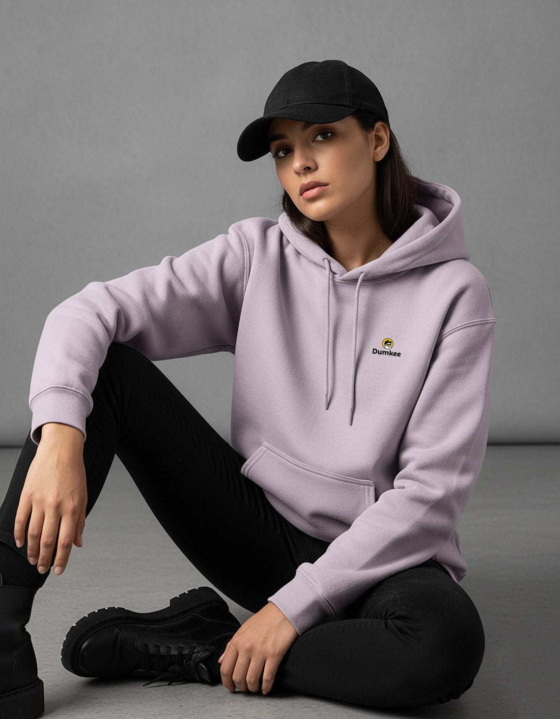 Person wearing a Dumkee baby pink hoodie with a logo, sitting on a gray floor.