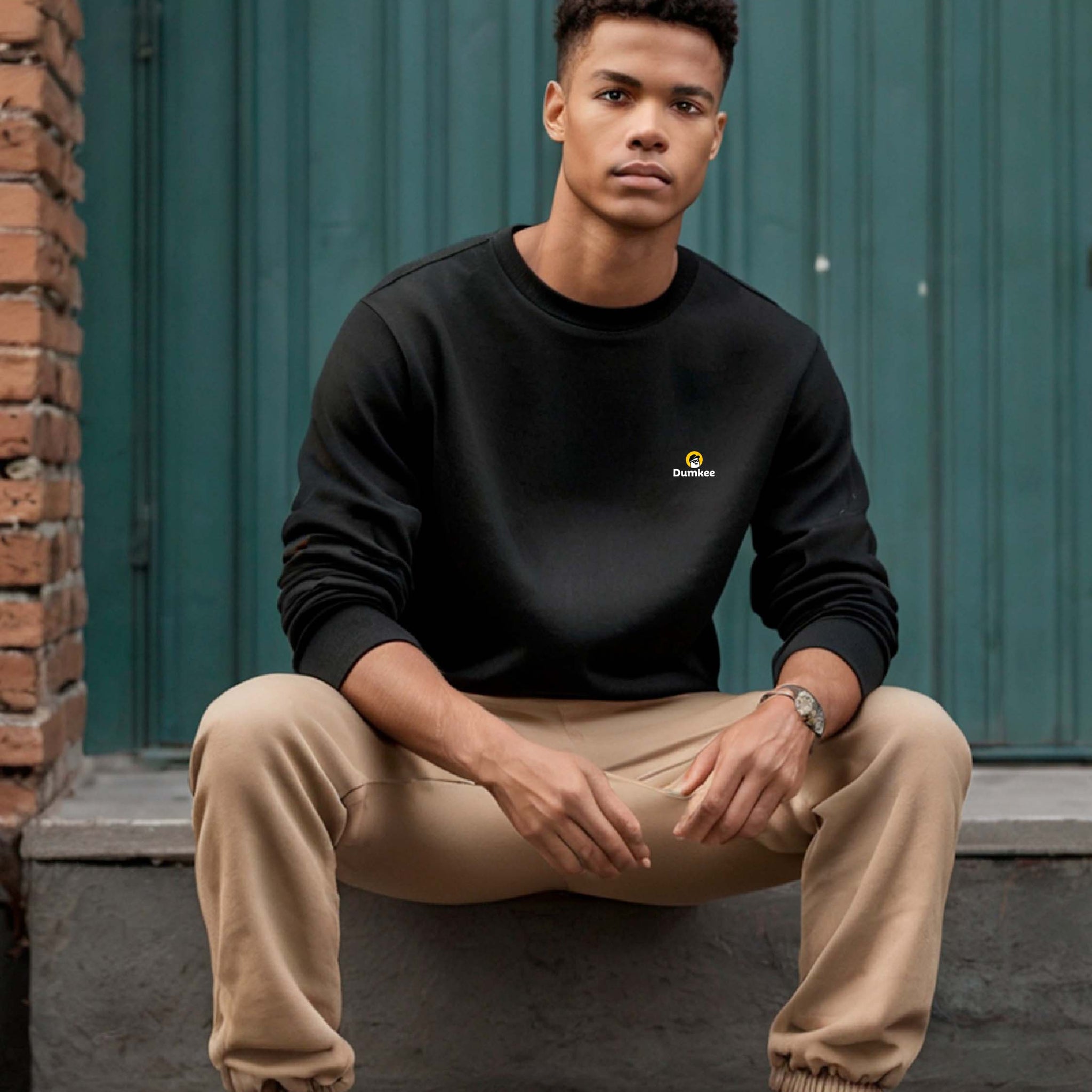 Man wearing a black sweatshirt with a Dumkee  logoand white sneakers sitting on a step.